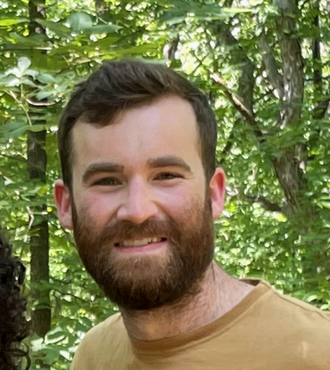 A smiling man with a beard and short dark hair, standing outdoors in a green wooded area.
