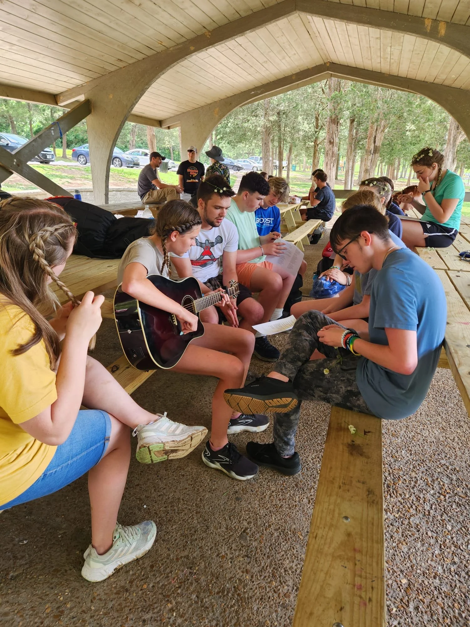 Group of young people sitting on wooden benches in a pavilion outdoors, some singing or reading, with one girl playing an acoustic guitar, surrounded by trees and parked cars in the background.
