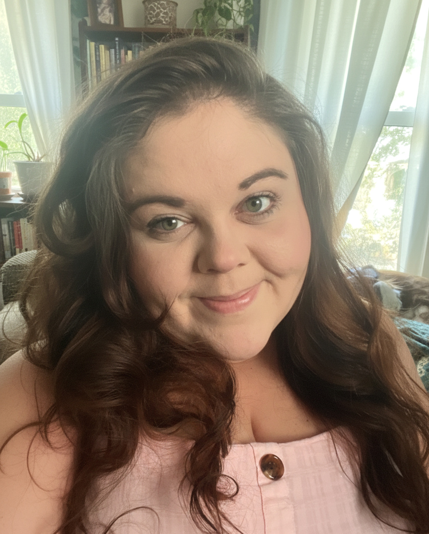 A woman with long, wavy brown hair and light skin smiling in a sunlit room with curtains, a bookshelf, and houseplants in the background.