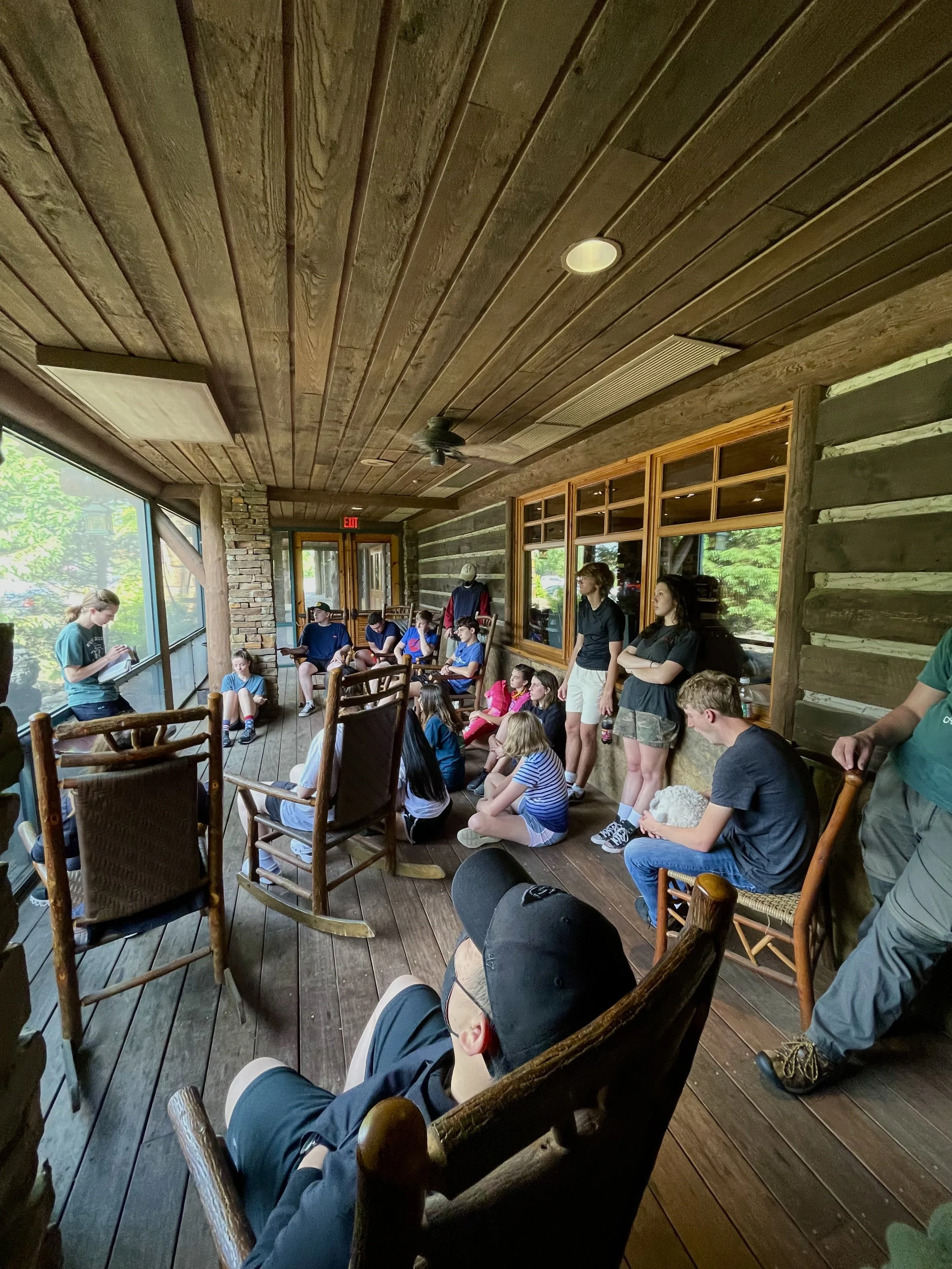 A group of children and teenagers sitting and standing in a rustic wooden room with large windows, listening to a woman reading from a notebook.