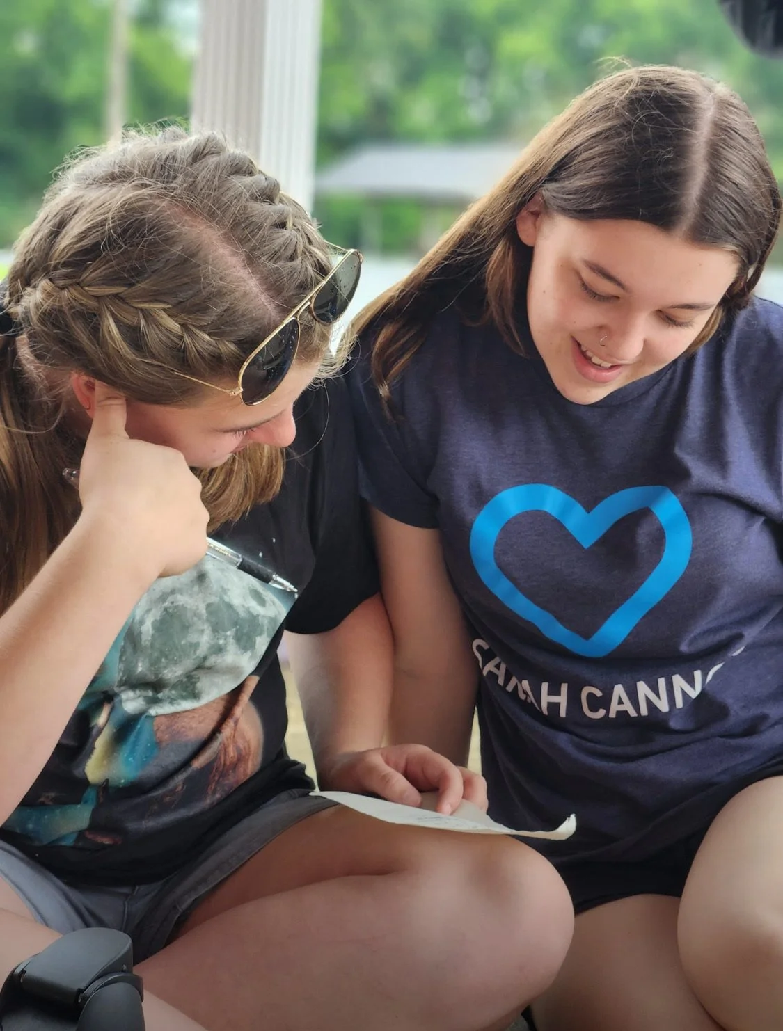 Two young women sitting outdoors, looking at a piece of paper together, one with sunglasses on her head and the other wearing a shirt with a blue heart and the words 'SETH CANN'.