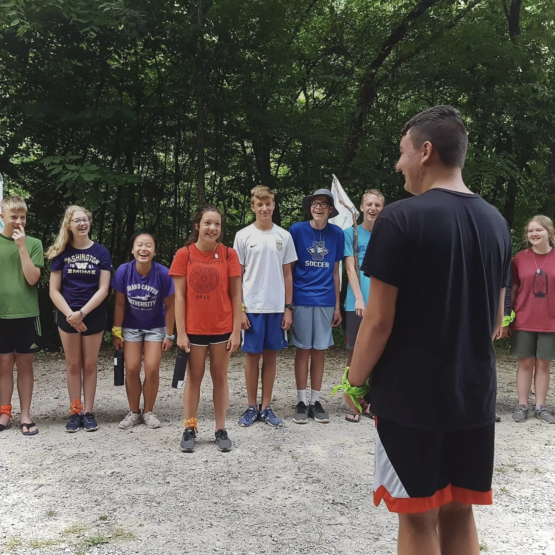 A group of children standing outdoors, laughing and smiling, as one boy faces them, holding a flag, on a gravel path surrounded by trees.