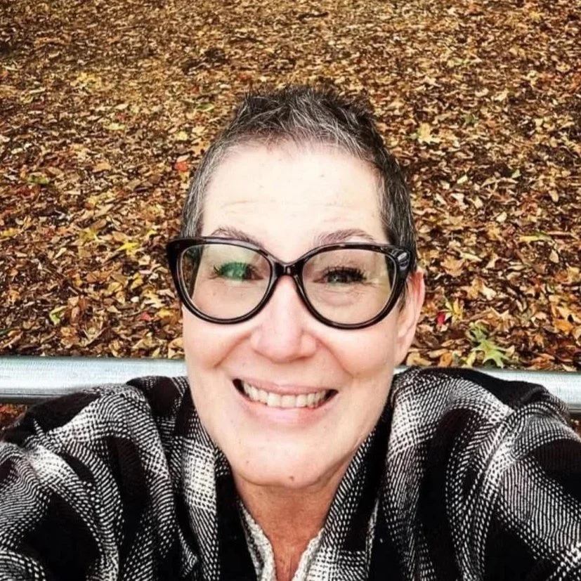 A person with short hair, wearing glasses and a black and white striped shirt, smiling for a selfie outdoors with fallen leaves on the ground.