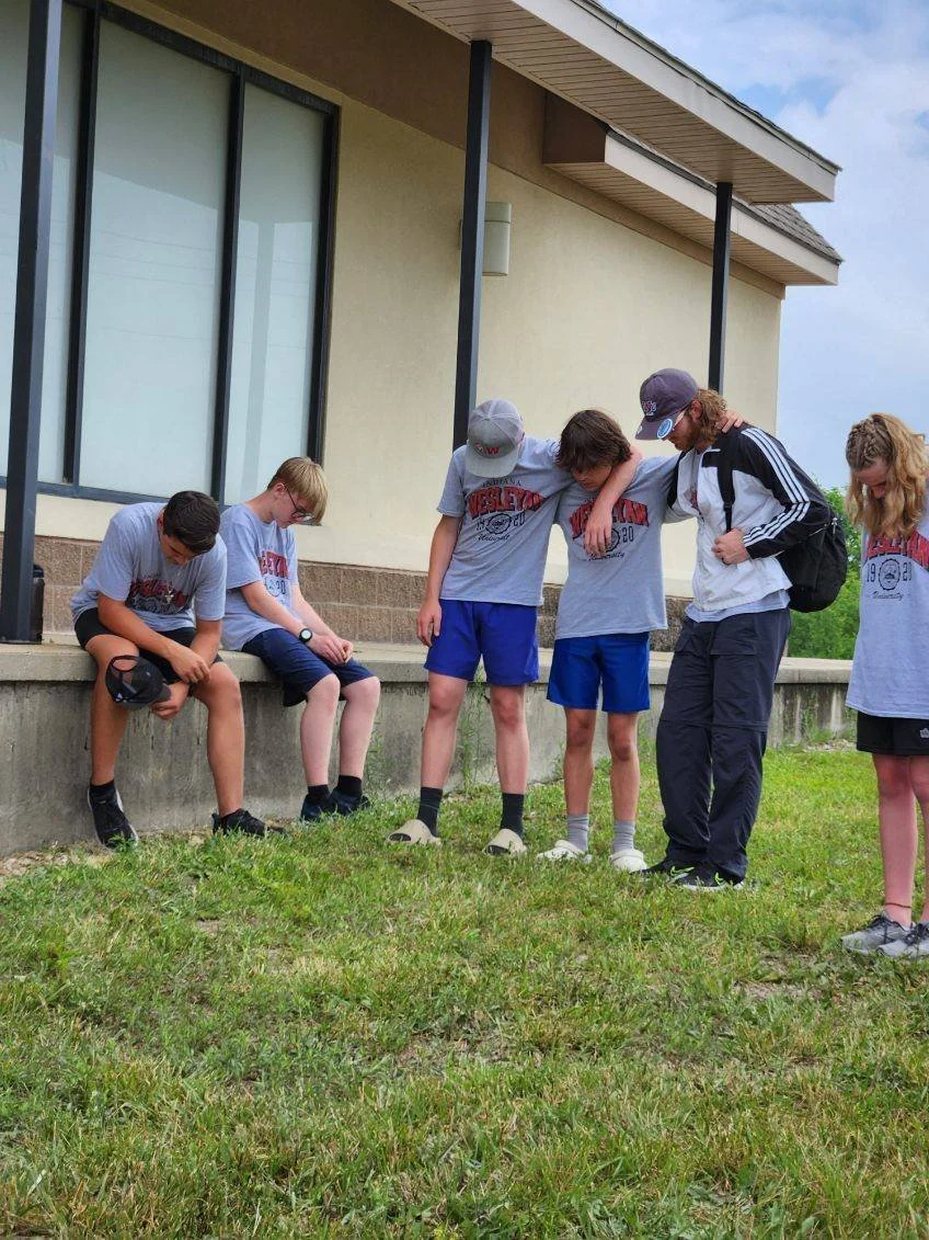 A group of young boys and a woman standing outdoors near a building, with some of the boys sitting and others standing with their heads bowed or leaning on each other, suggesting a moment of support or reflection.