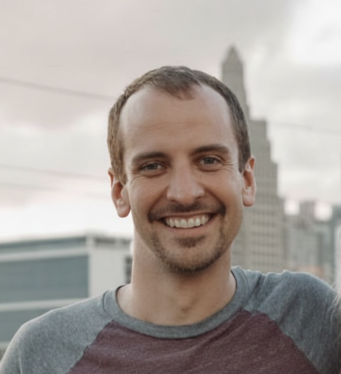 Smiling man outdoors with city skyline in the background, wearing a gray and maroon shirt.