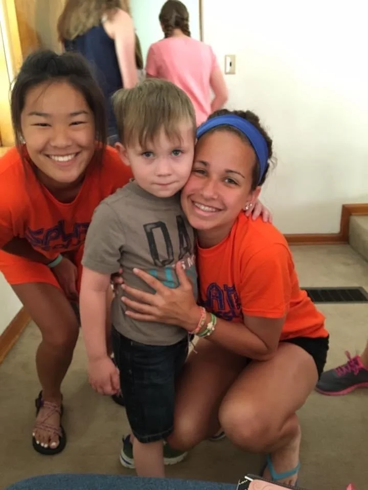 Three children smiling and hugging each other indoors. One young girl with dark hair and a bright orange shirt, a young boy with light brown hair and a gray T-shirt, and another girl with dark curly hair, a blue headband, and an orange shirt.