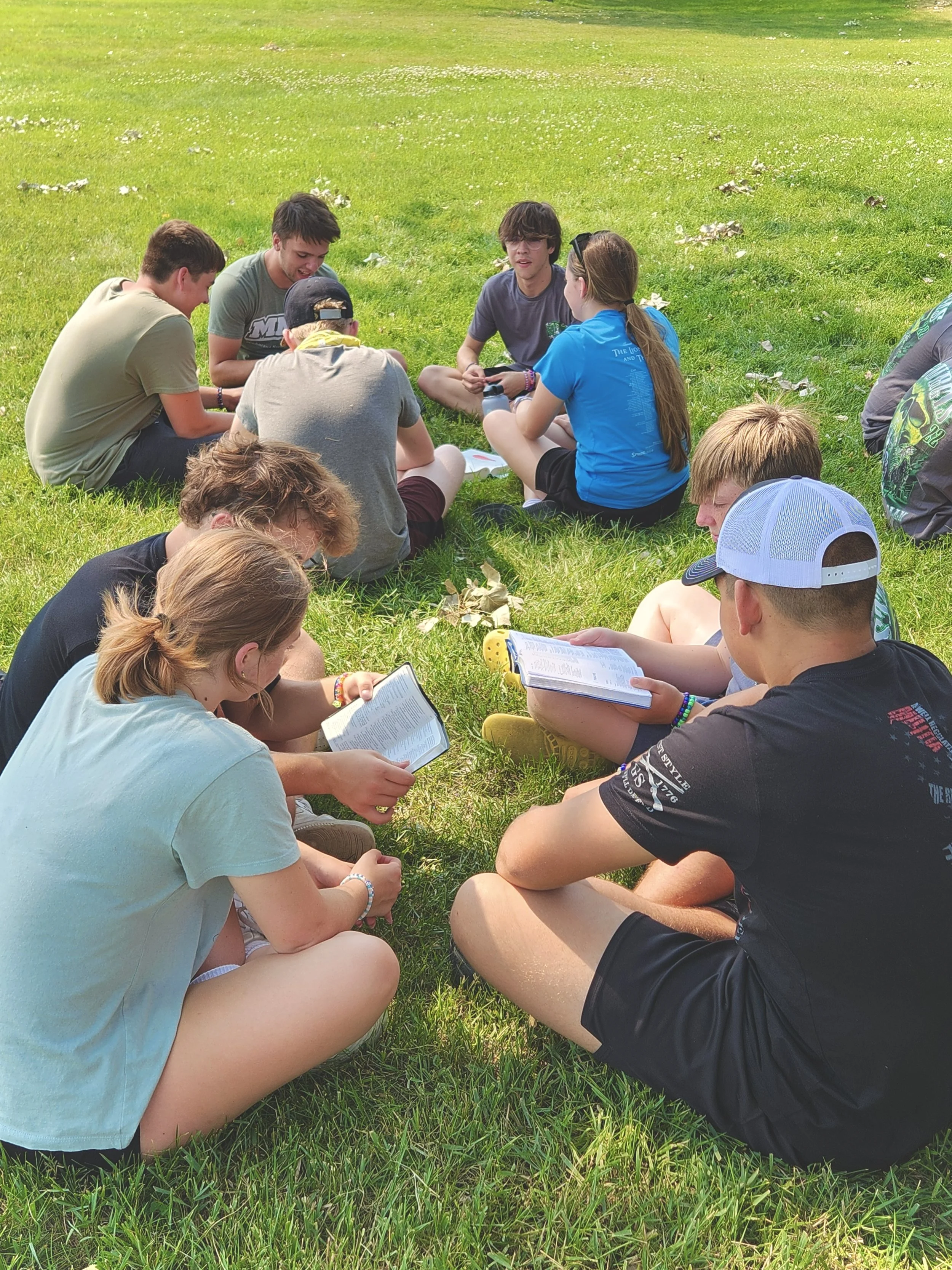 A group of teenagers sitting on grass in a circle, some reading books and others talking, outdoors on a sunny day.