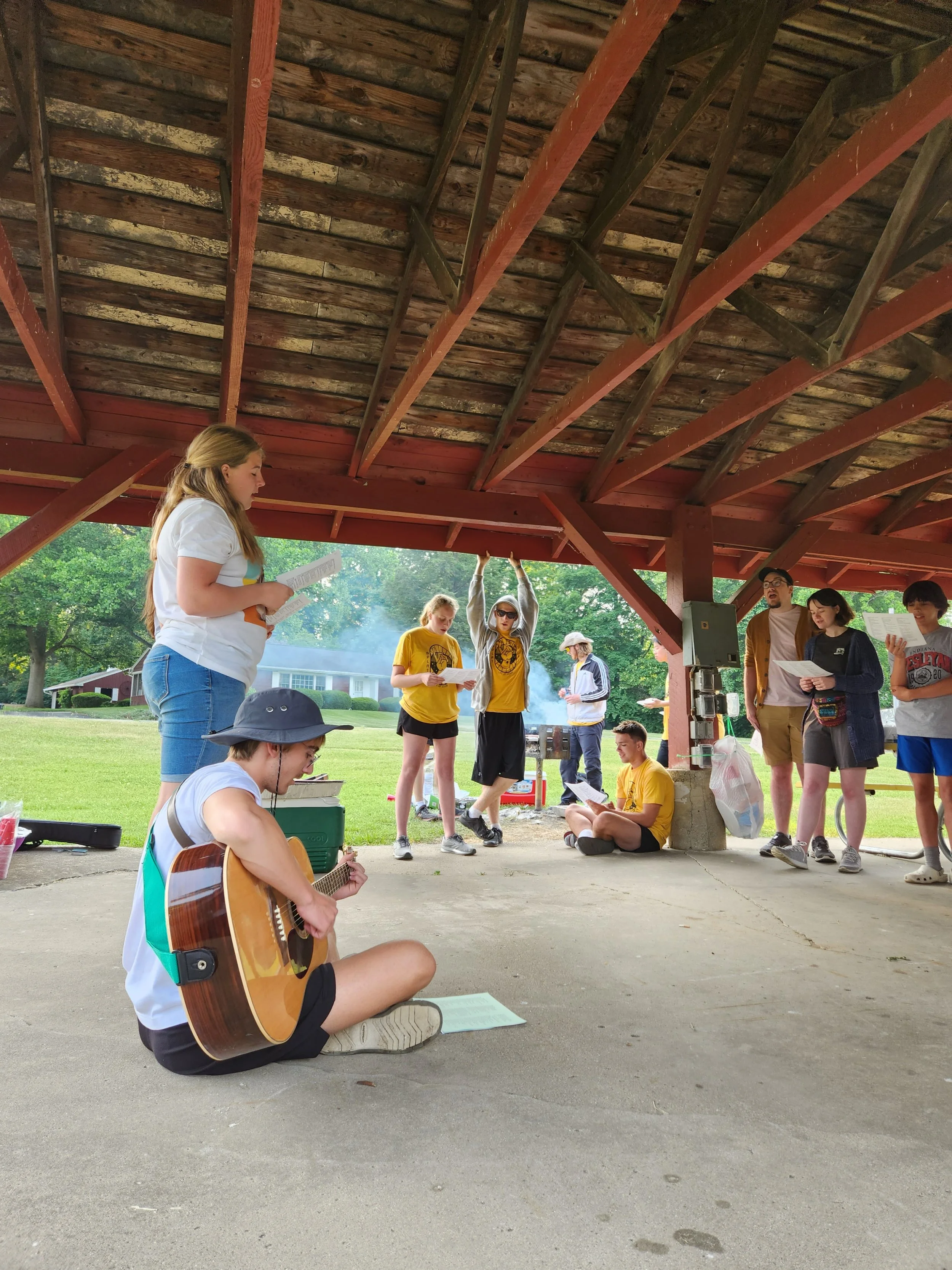Group of young people gathered under a pavilion in a park, some singing, some playing guitar, and others reading, with trees and a house in the background.
