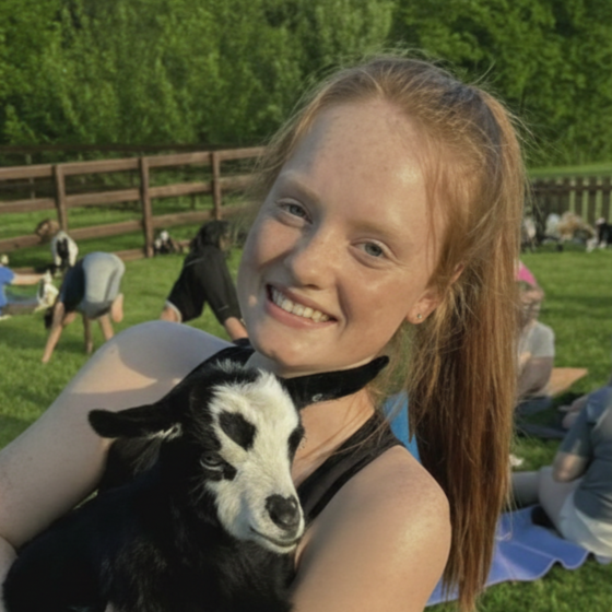 A red-haired girl smiling and holding a black and white goat kid outdoors on a sunny day, with a wooden fence, green grass, and trees in the background.