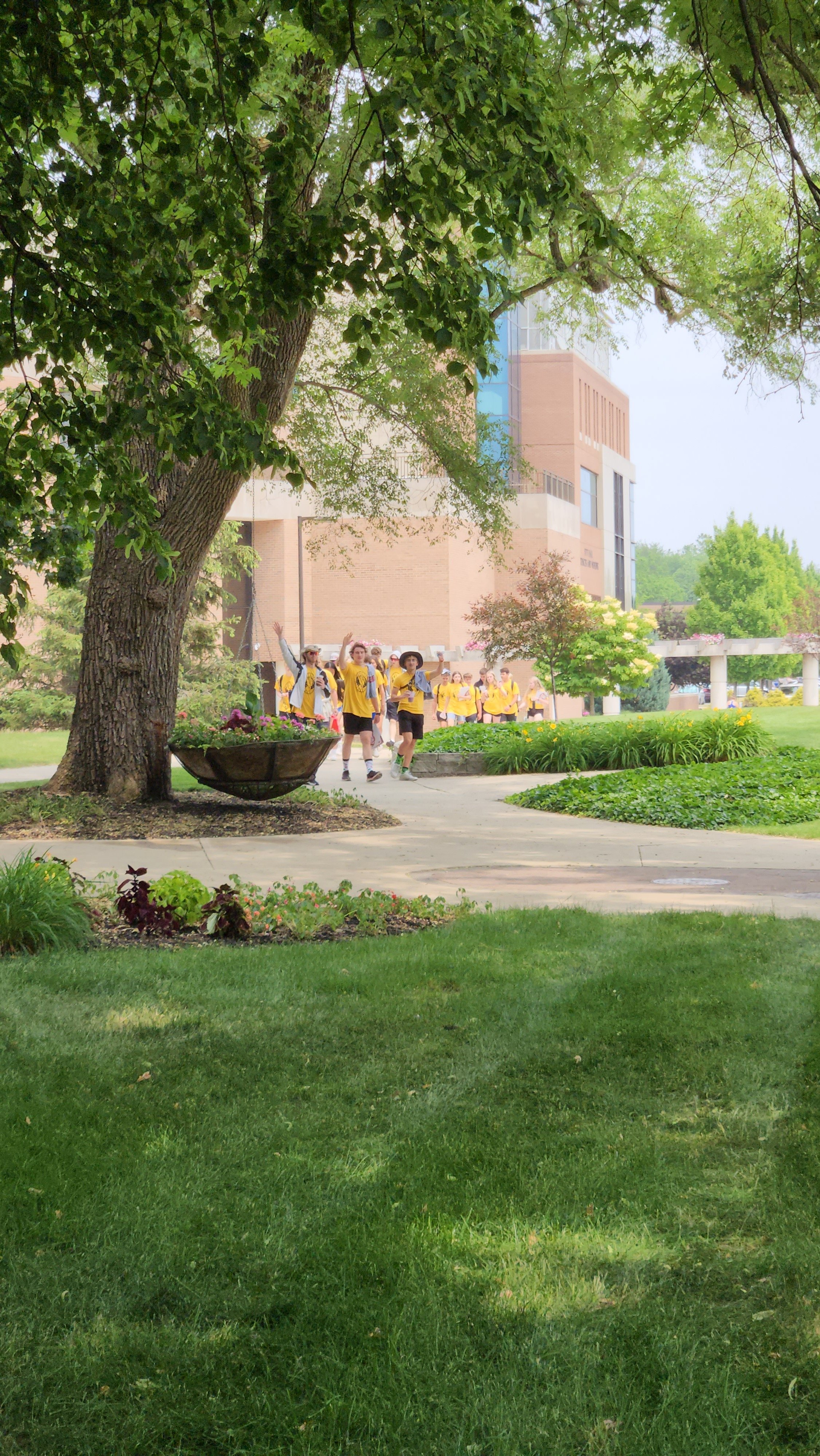 A group of children wearing yellow shirts and backpacks, walking through a park-like area with green grass, trees, and flowers on a sidewalk, with a large building in the background.
