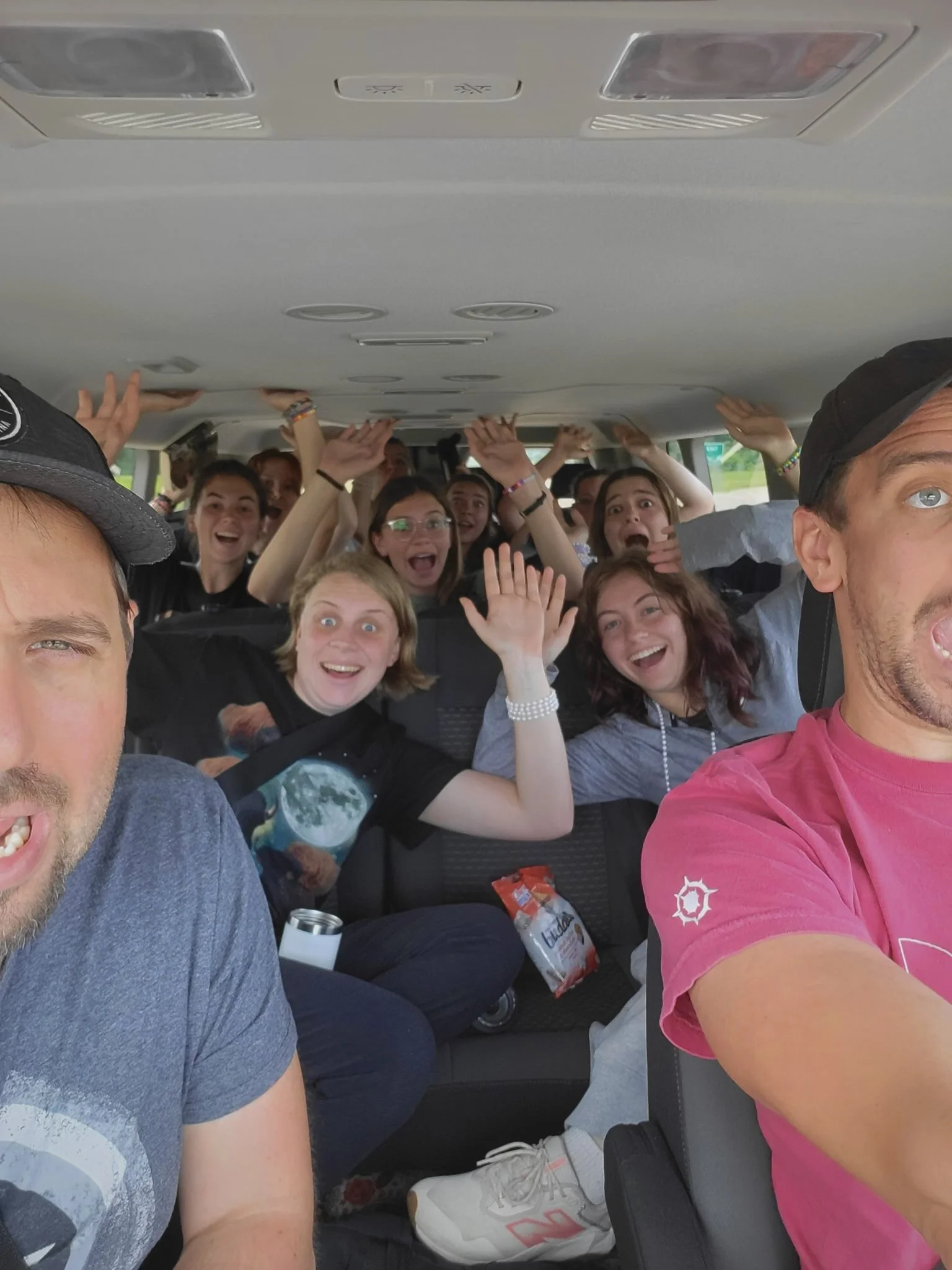 Group of smiling young people and two men taking a selfie inside a vehicle, with some raising their hands and making gestures.