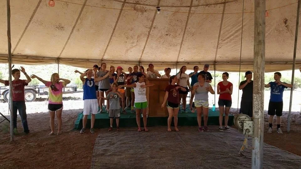 Group of children and teenagers standing under a large tent, some with their arms raised or crossed, appearing to participate in an activity or performance on a small stage.