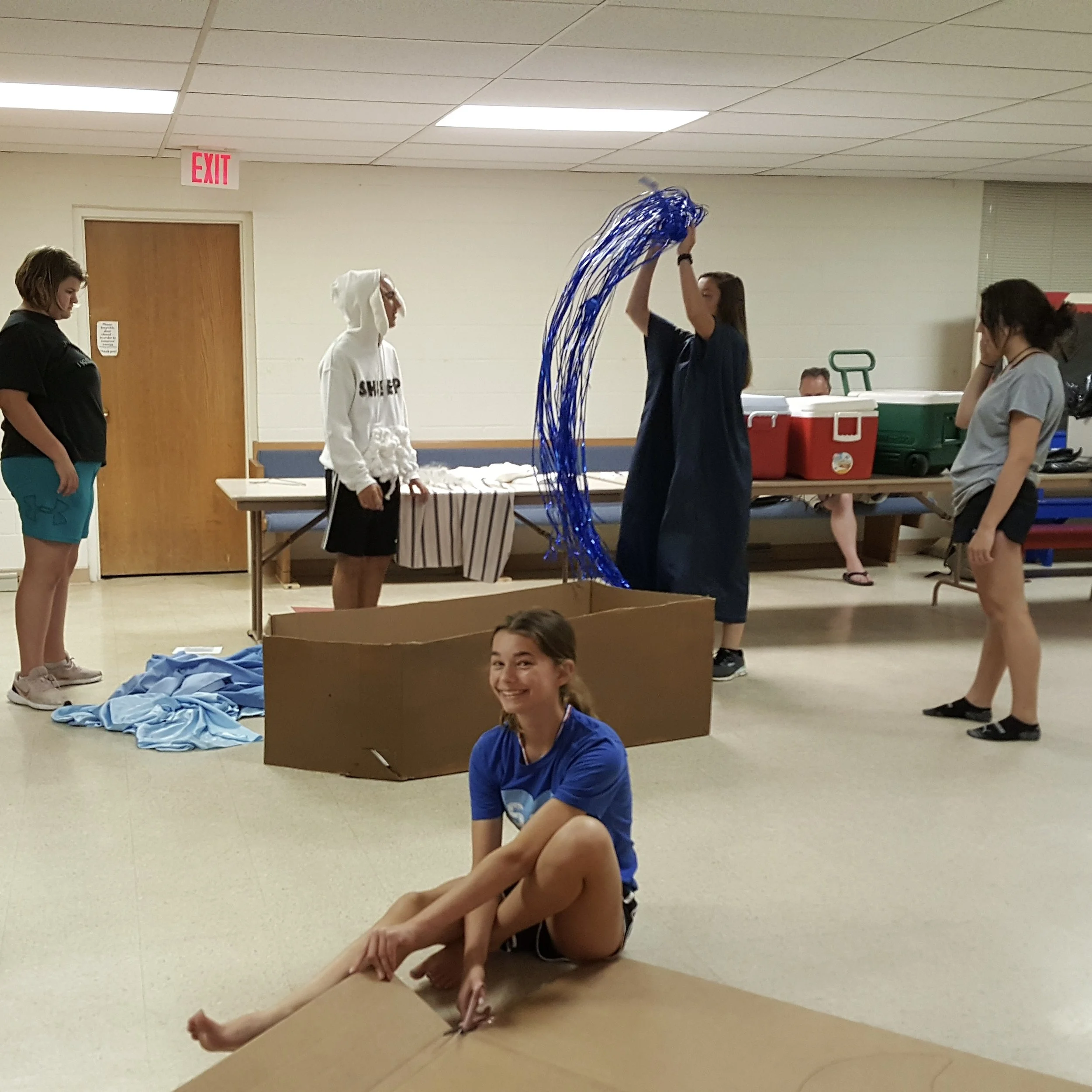 Young girl smiling and sitting on the floor, surrounded by other children in a room with a decorated setup, possibly for a school or community event.