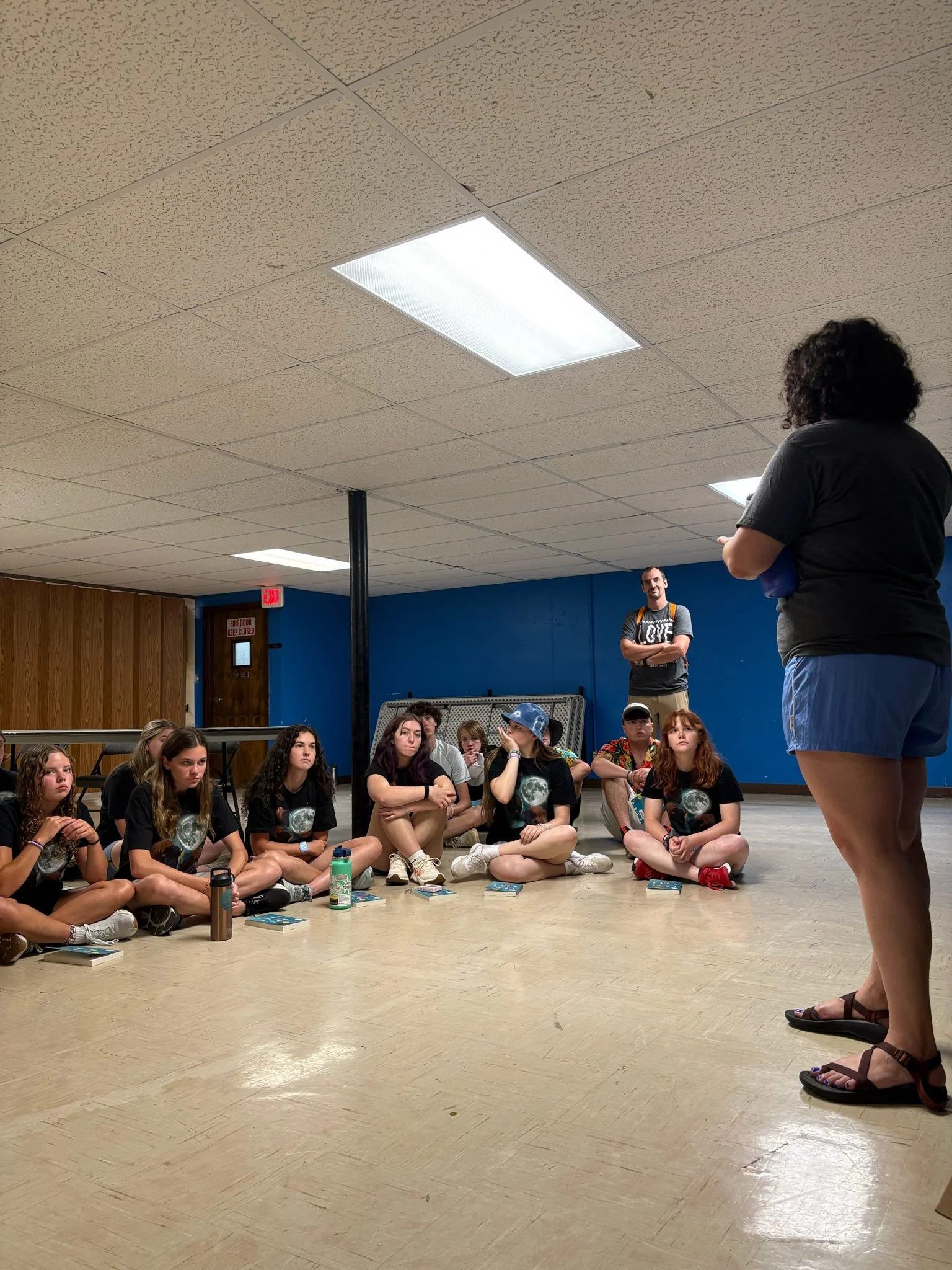 A group of young girls sitting on the floor in a circle, listening to a woman standing in front of them, inside a room with a blue wall and ceiling lights.