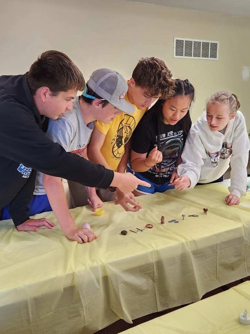 Five children gathered around a table, closely examining small objects laid out on a yellow tablecloth.