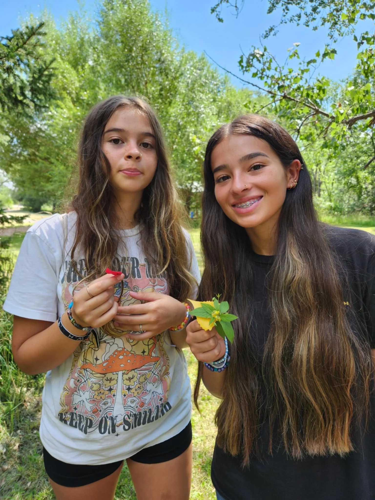 Two young girls standing outdoors in a lush green park, smiling and holding colorful leaves and small objects, with trees and a blue sky in the background.
