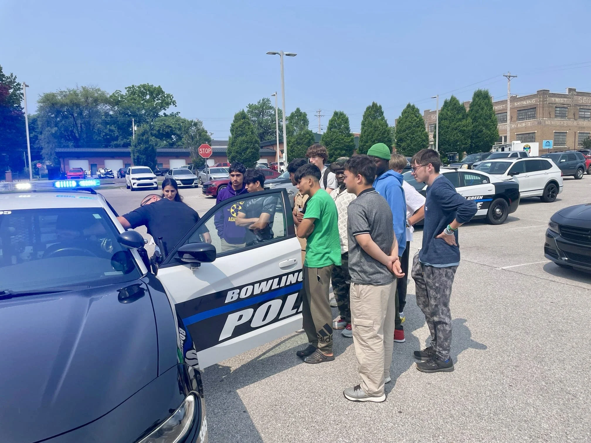 A group of teenagers gathered around a police car in a parking lot, listening to a police officer during a community event.