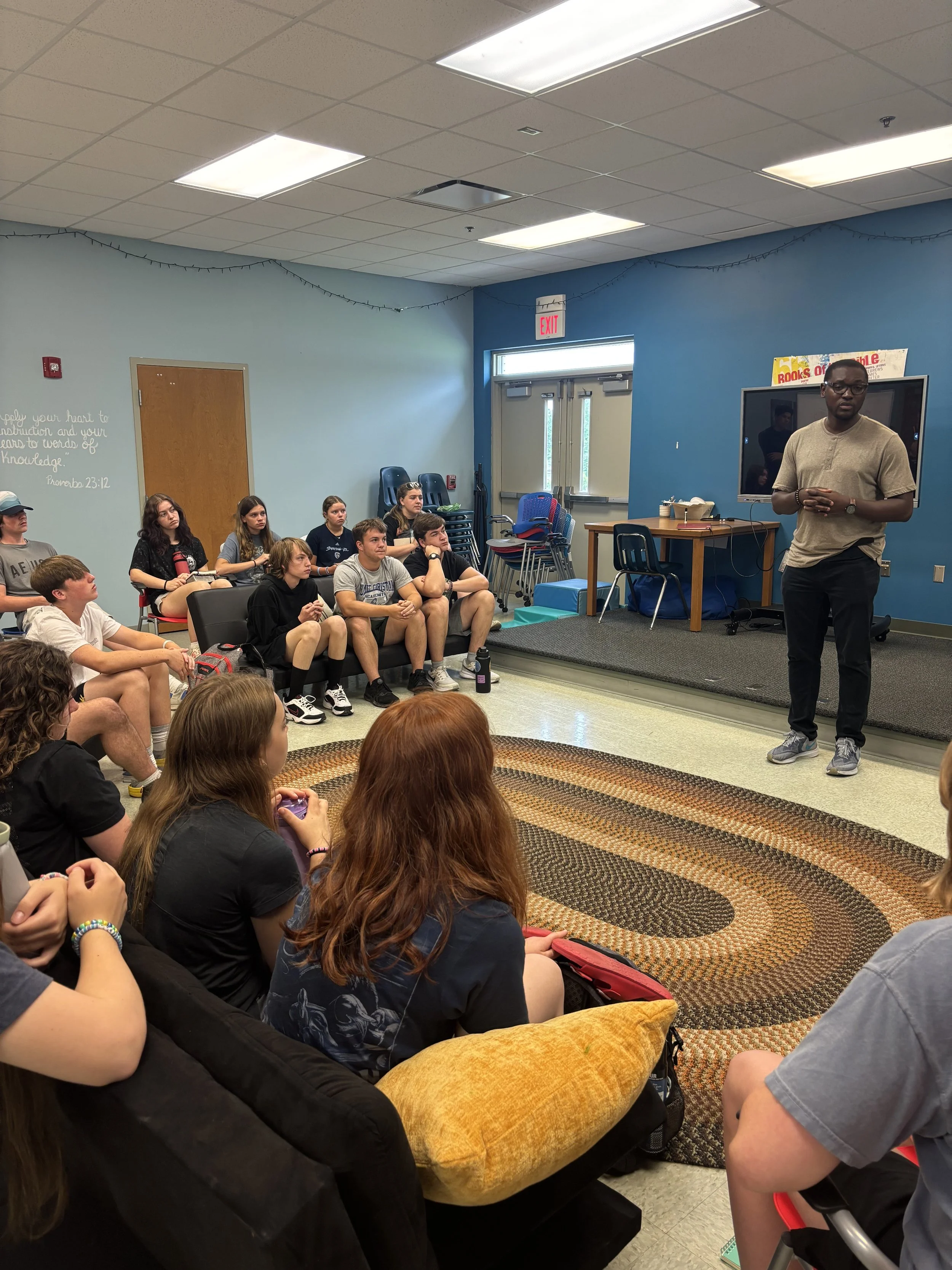 A group of students sitting on chairs and couches in a classroom or meeting room, attentively listening to a man standing in front near a large screen.