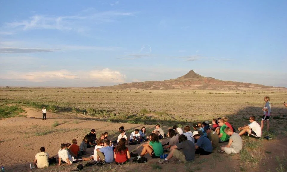 A group of people sitting outdoors on the desert ground, listening to a person standing nearby with a vast desert landscape and mountain in the background.