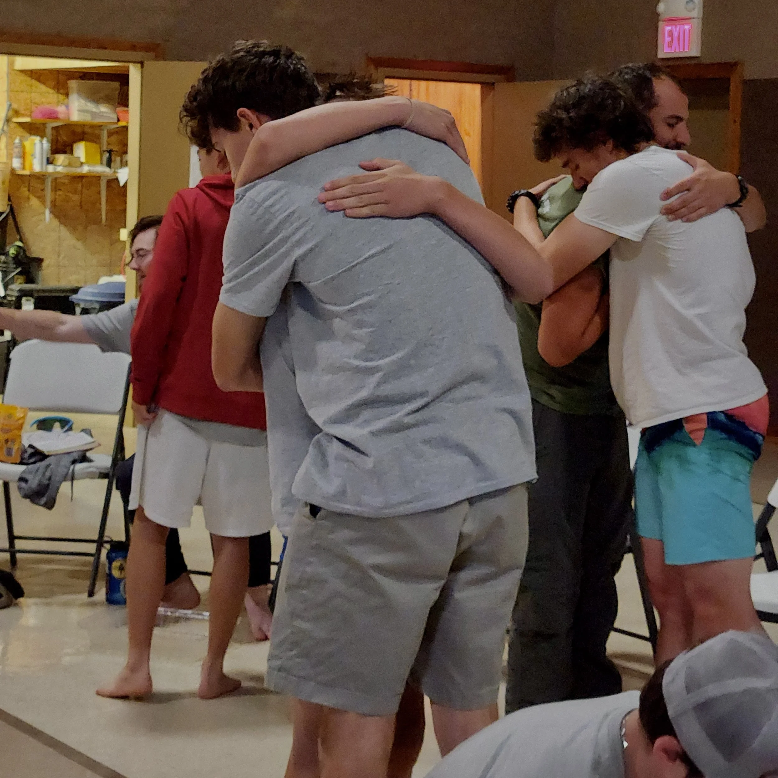 Group of young people hugging in a circle indoors, some standing barefoot, with casual clothing and relaxed atmosphere.