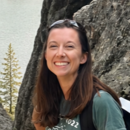 A woman with brown hair, smiling and showing her teeth, wearing a maroon shirt outdoors near water with a blurred background.