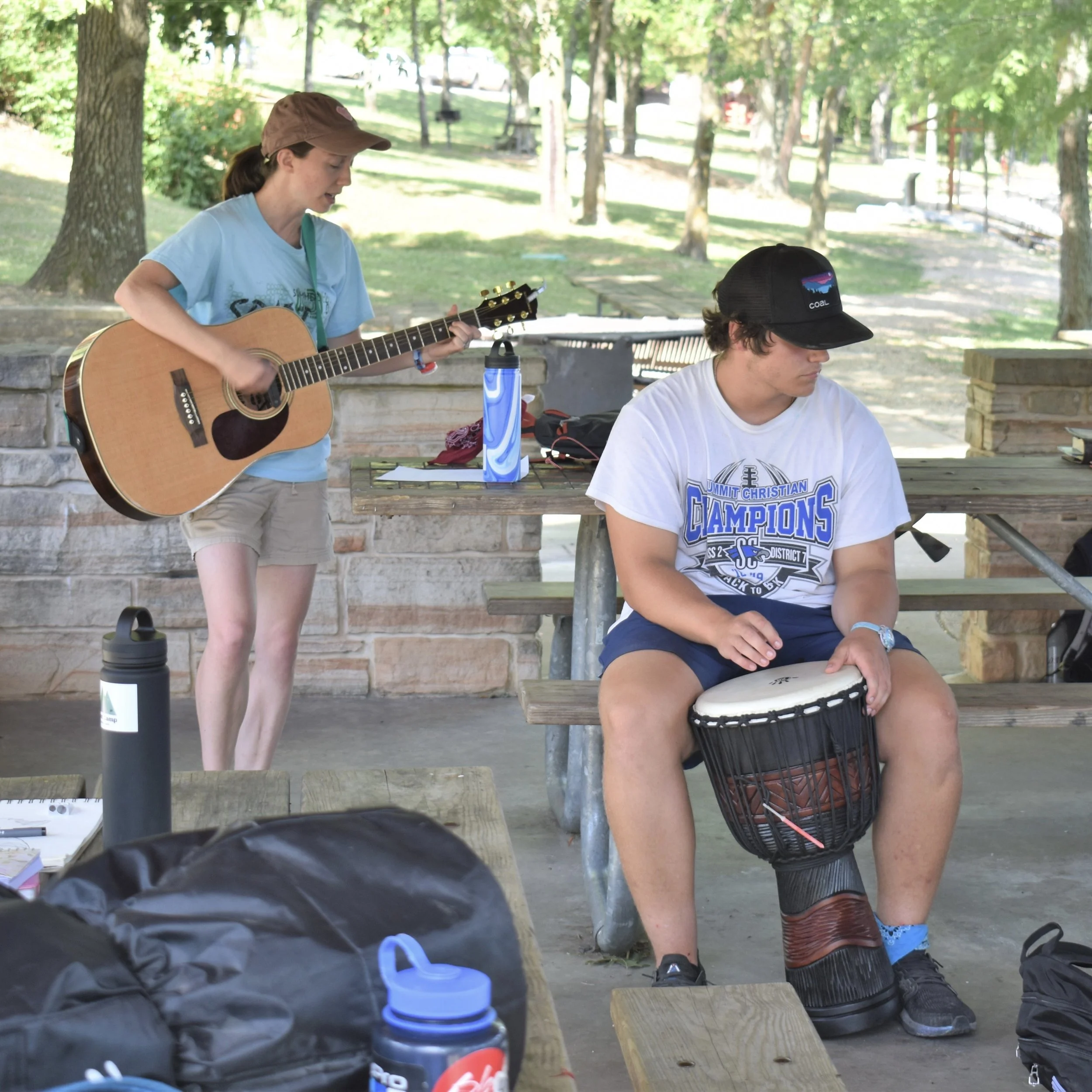 A girl playing an acoustic guitar and a boy playing a drum at an outdoor park with trees, picnic tables, and a stone wall in the background.