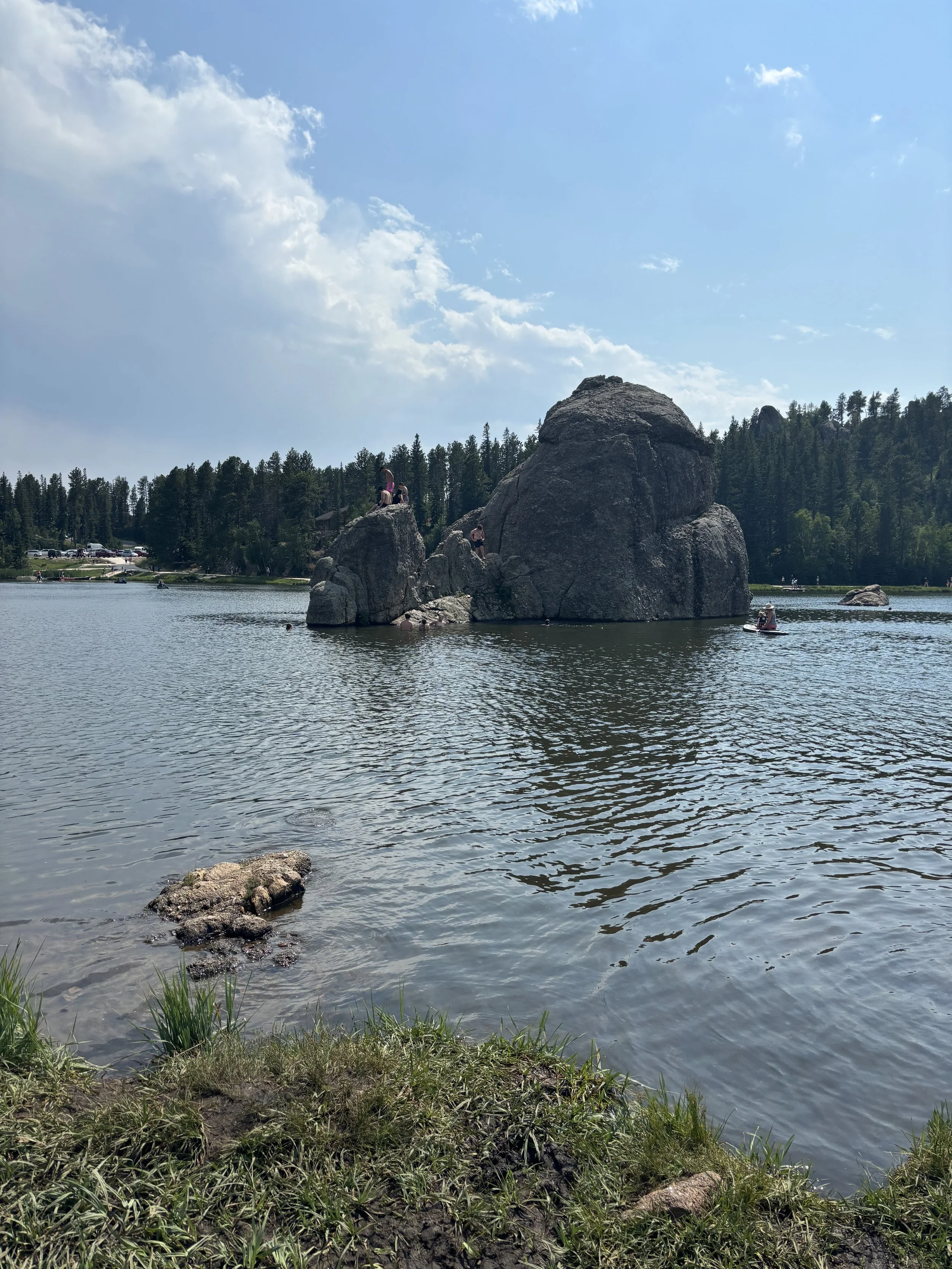 A lake with large rocks in the water, surrounded by forested land under a partly cloudy sky, with some people on the rocks and in a boat.