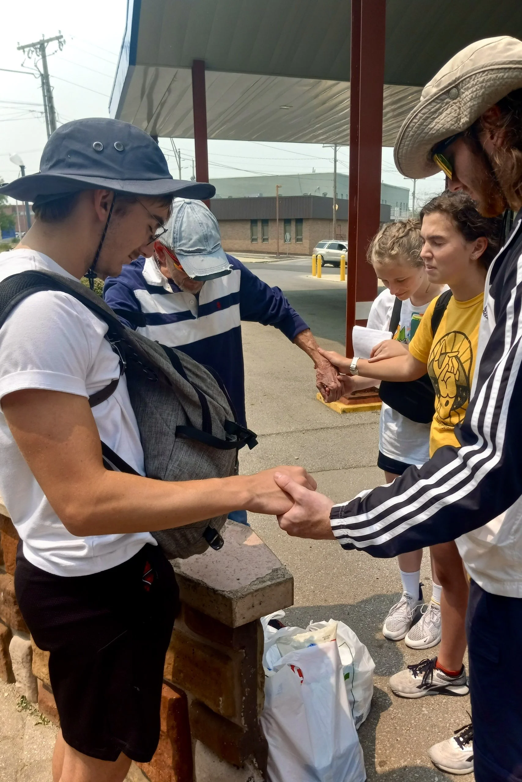 People holding hands in a prayer or moment of silence outside under a shelter, with brick and concrete surroundings and a street in the background.