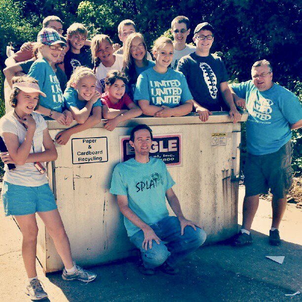 Group of children and adults gathered around a large paper and cardboard recycling bin outdoors on a sunny day.