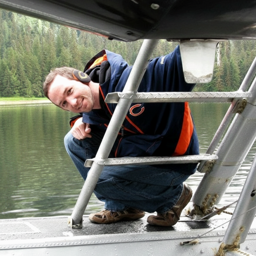 Man crouching on the deck of a boat near a lake, smiling at the camera with a forested shoreline in the background.