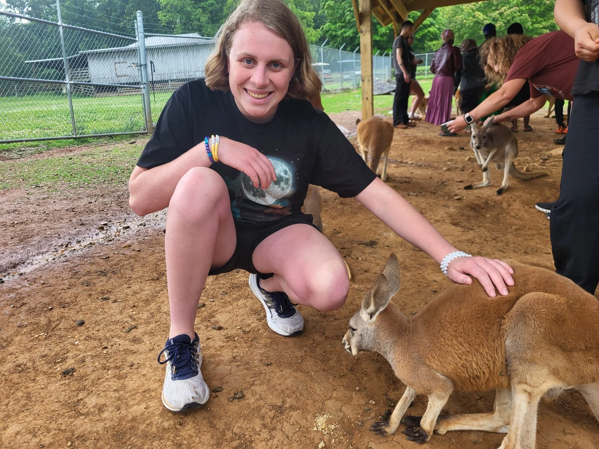 A young woman smiling and squatting on the ground in a petting zoo, petting a small kangaroo while other people and kangaroos are visible in the background.