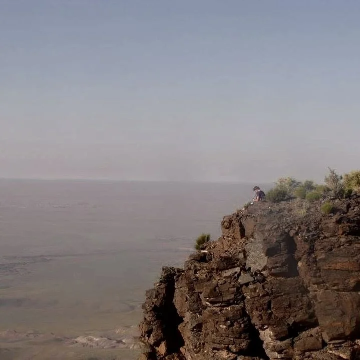 Person sitting on a rocky cliff overlooking the ocean on a clear day with a light blue sky.