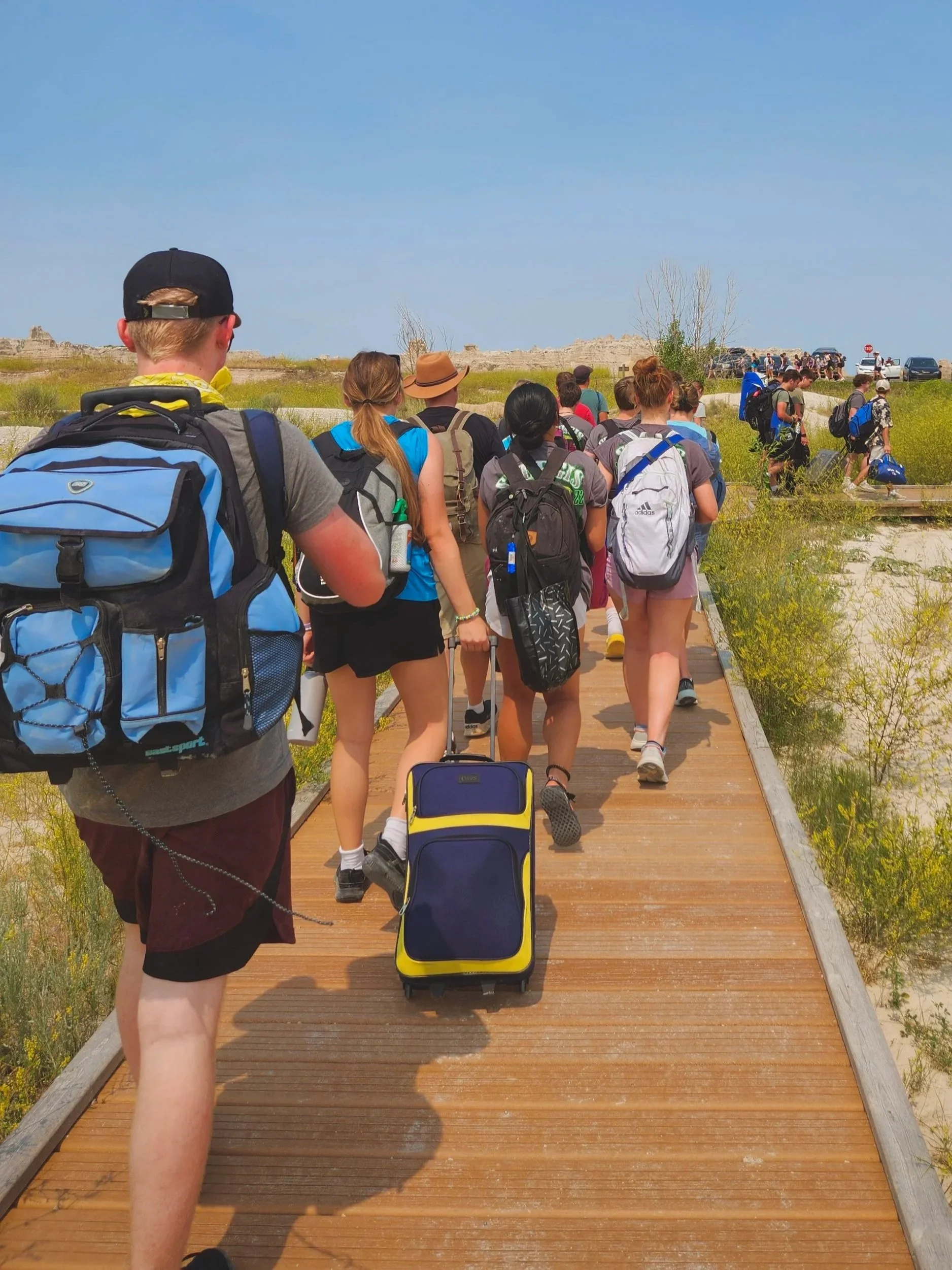 Group of people walking on a wooden boardwalk outdoors in a sunny area with grass and open landscape.