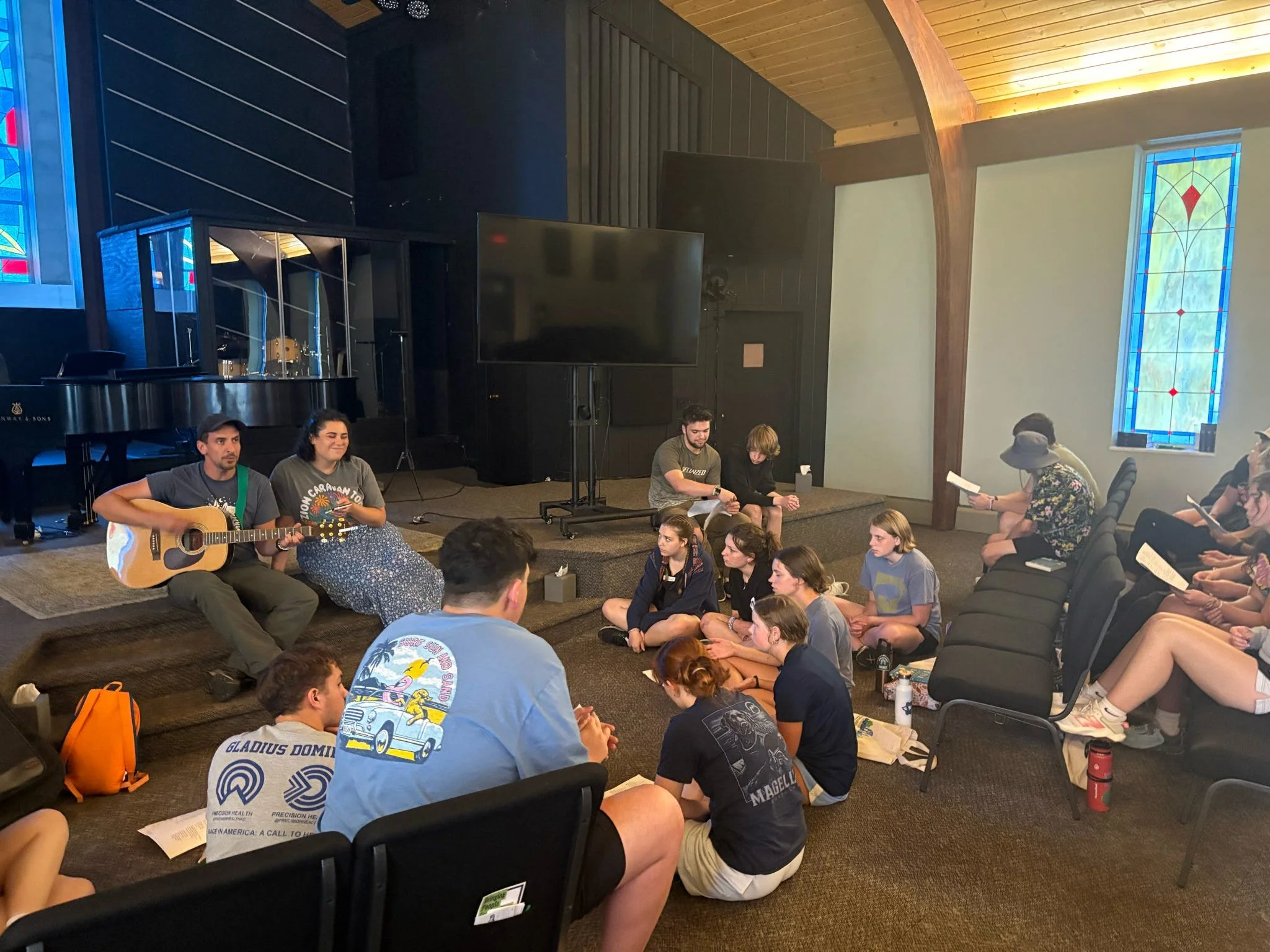 A group of young people gathered in a church or community room, sitting on the floor and chairs, listening to two musicians performing with a guitar and a tambourine on a small stage. Some are following along with songbooks, while others look attenti