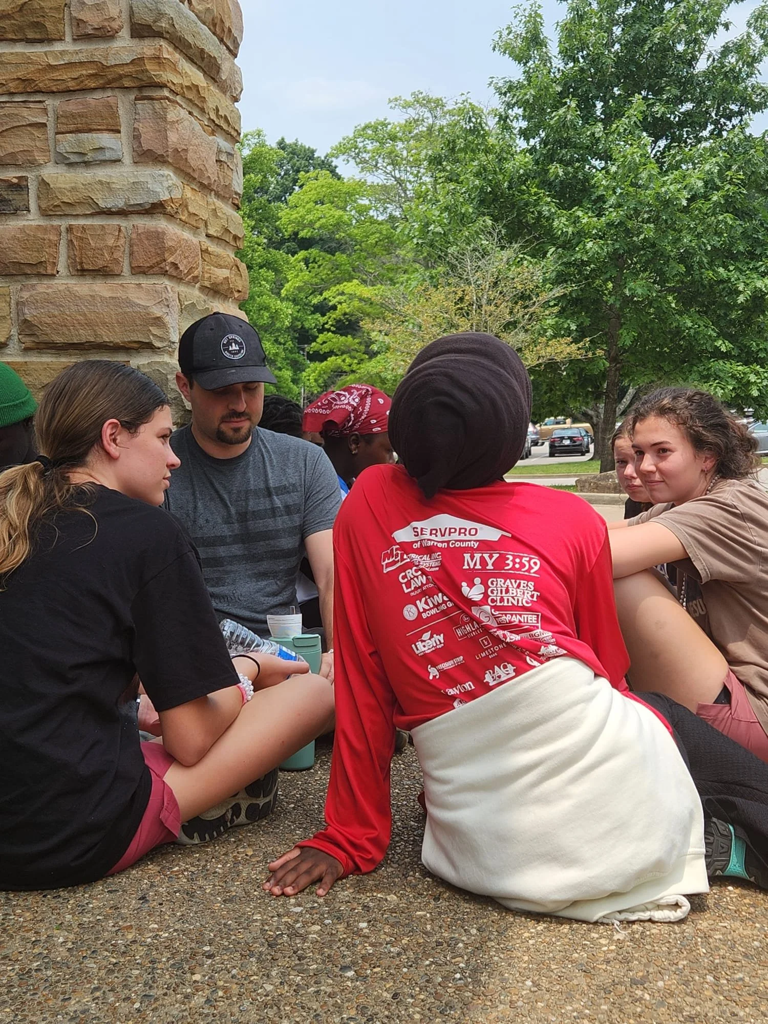 Group of young people sitting outdoors on a sidewalk, engaging in conversation; background features green trees and a brick wall.