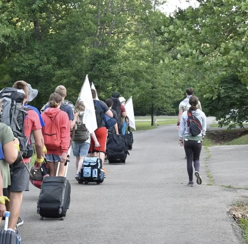 Group of young people walking with rolling suitcases and backpacks on a paved path through a green park, some holding white flags.