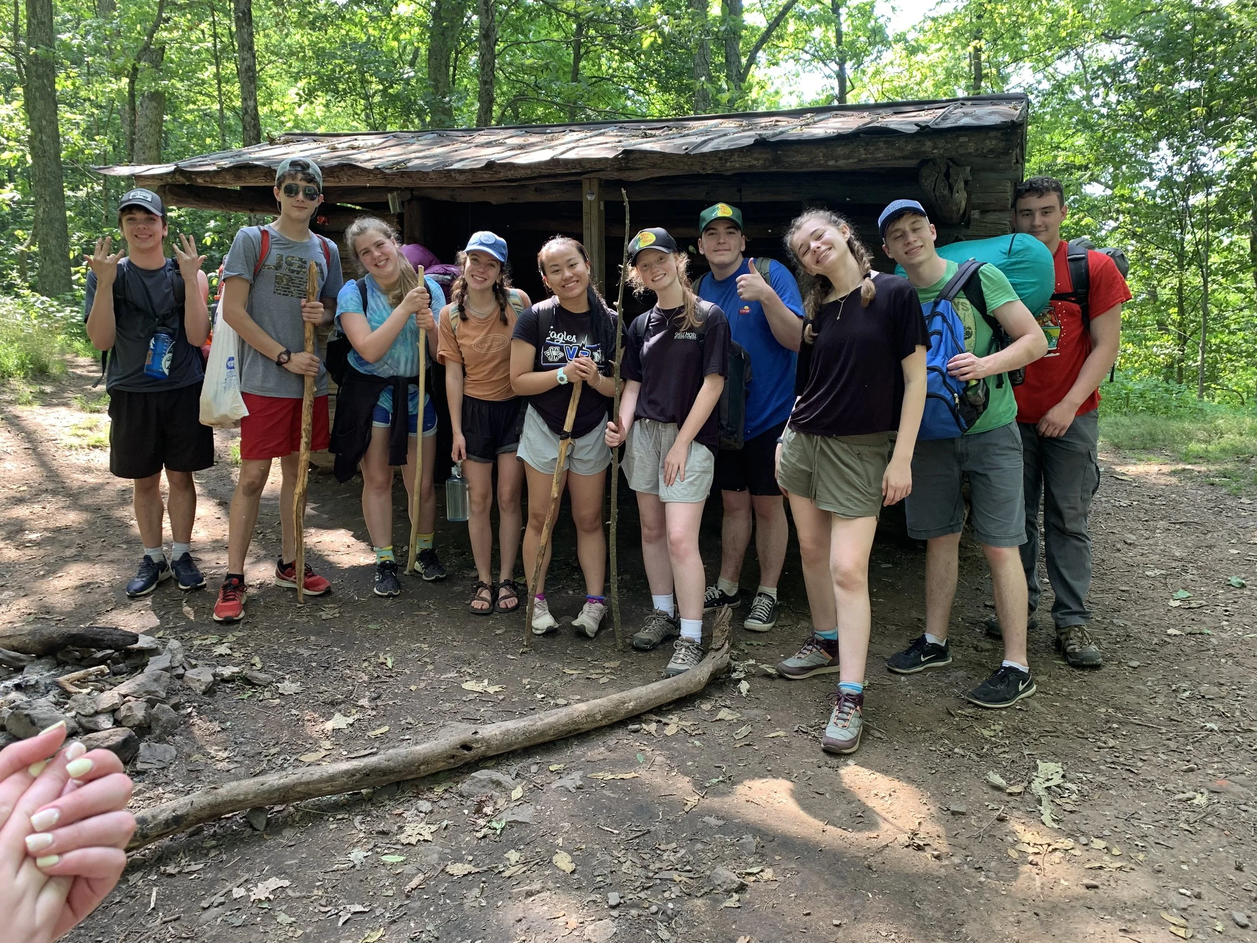 Group of young people standing in front of a small wooden shelter in a wooded area during a hiking trip, some holding trekking sticks, wearing backpacks and outdoor clothing.