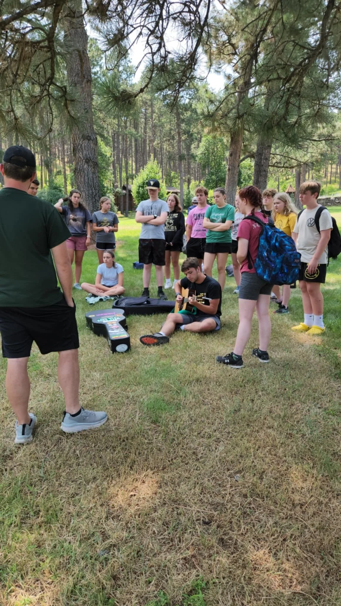 A group of young people gathered outdoors under tall trees, with one person sitting on the grass playing guitar while others stand around listening. An individual with a black cap and black shorts is in the foreground.