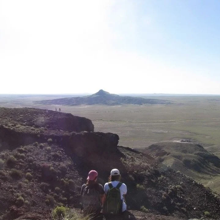 Two hikers with backpacks and hats sitting on a rocky hillside overlooking a vast, flat landscape with a distant mountain formation under a clear blue sky.