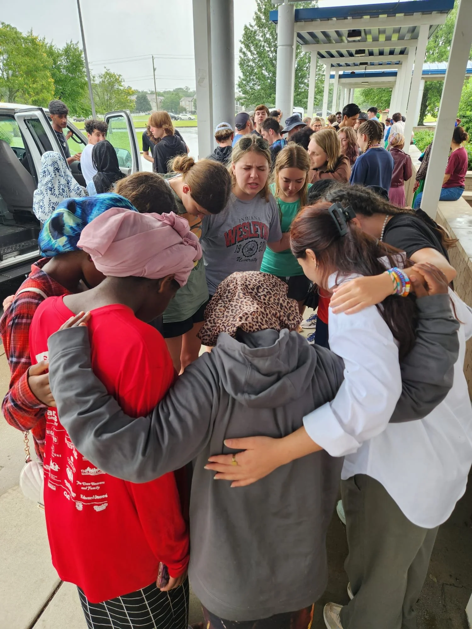 A diverse group of people gathered outdoors under a covered area, with some people in the foreground praying or holding hands in a circle, and others standing nearby or waiting.