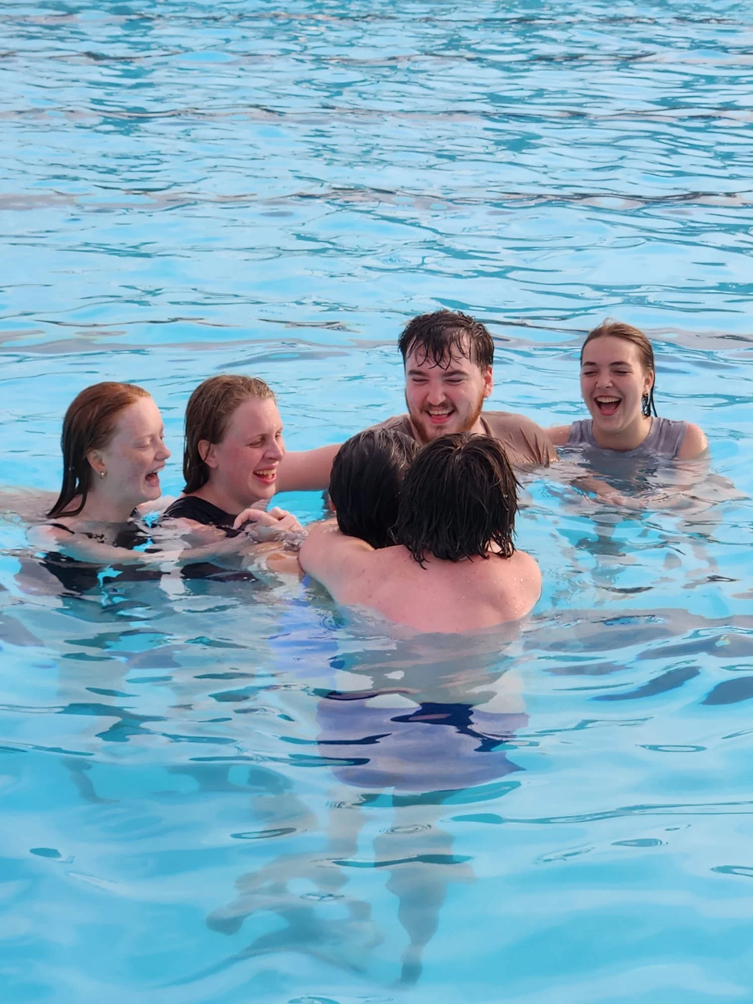 Group of five young adults joyfully hugging and laughing while swimming in a clear blue pool.