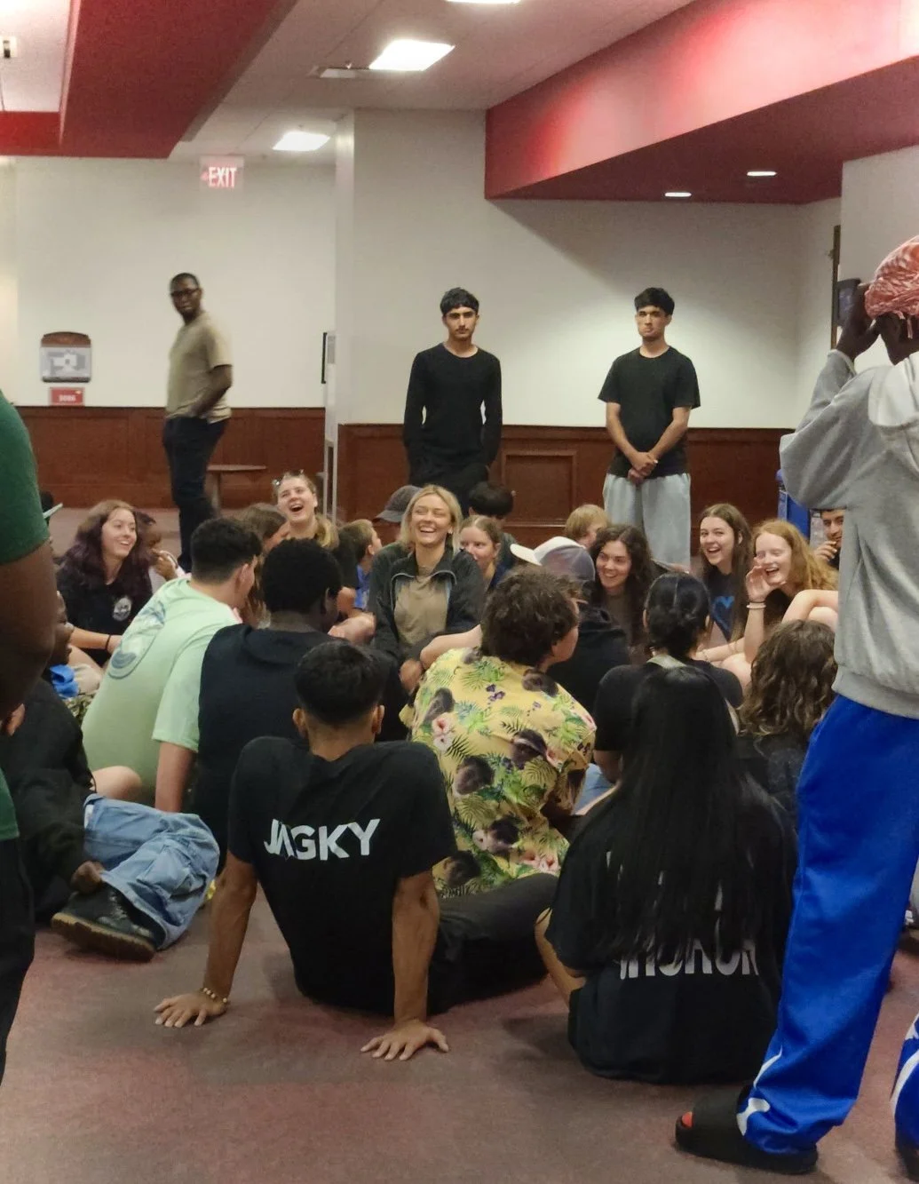 Group of teenagers sitting on the floor and laughing at an indoor gathering. Two boys stand behind them against the wall. The room has wood paneling and a red ceiling.