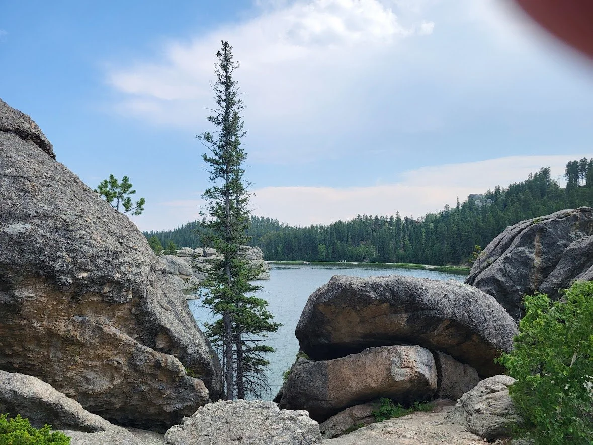A serene lakeside scene with large rocks and boulders in the foreground, tall pine trees, and a calm lake with a forested shoreline under a partly cloudy sky.