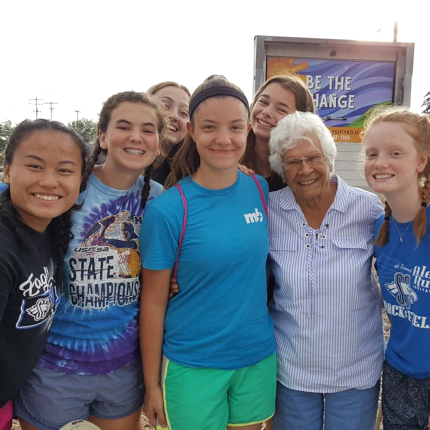 Group of young girls and an elderly woman smiling outdoors, with a sign in the background that says "Be the Change."