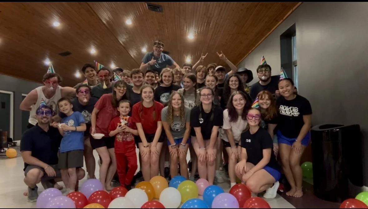 Group of people celebrating a party with balloons and party hats in an indoor room with wooden ceiling.