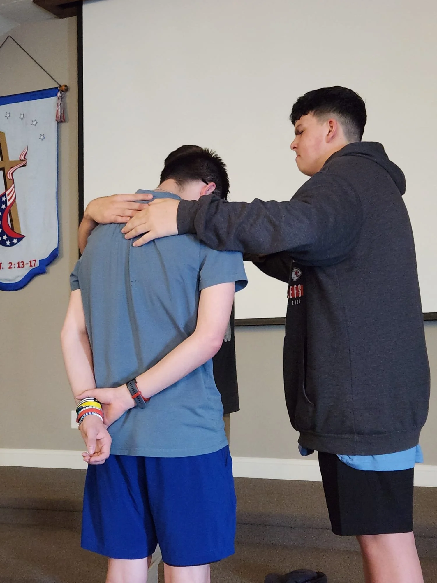A young boy with hands clasped behind his back stands with his head bowed as a man places his hand on the boy's head in a gesture of prayer or blessing. The setting appears to be indoors in a room with a plain wall and a banner partially visible on t
