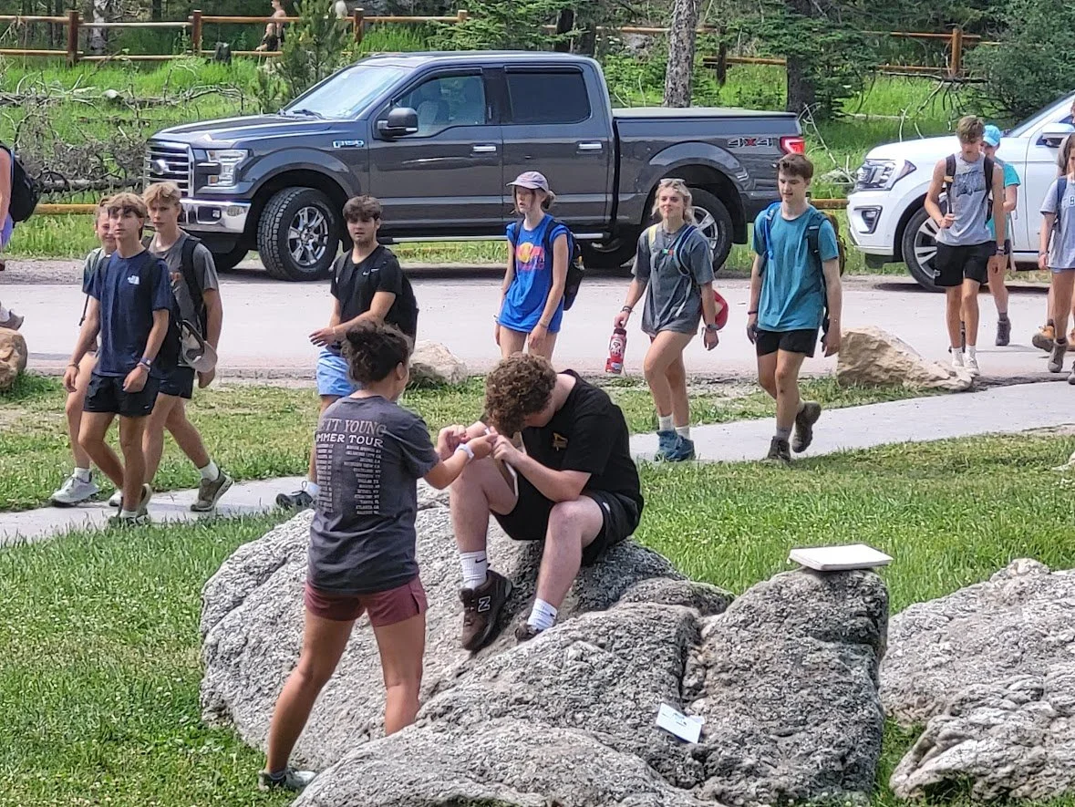 A group of children walking in a park with a man sitting on a large rock. The children are wearing backpacks and casual clothes, and the man appears to be engaged with a girl in front of him. There are parked cars and a wooden fence in the background