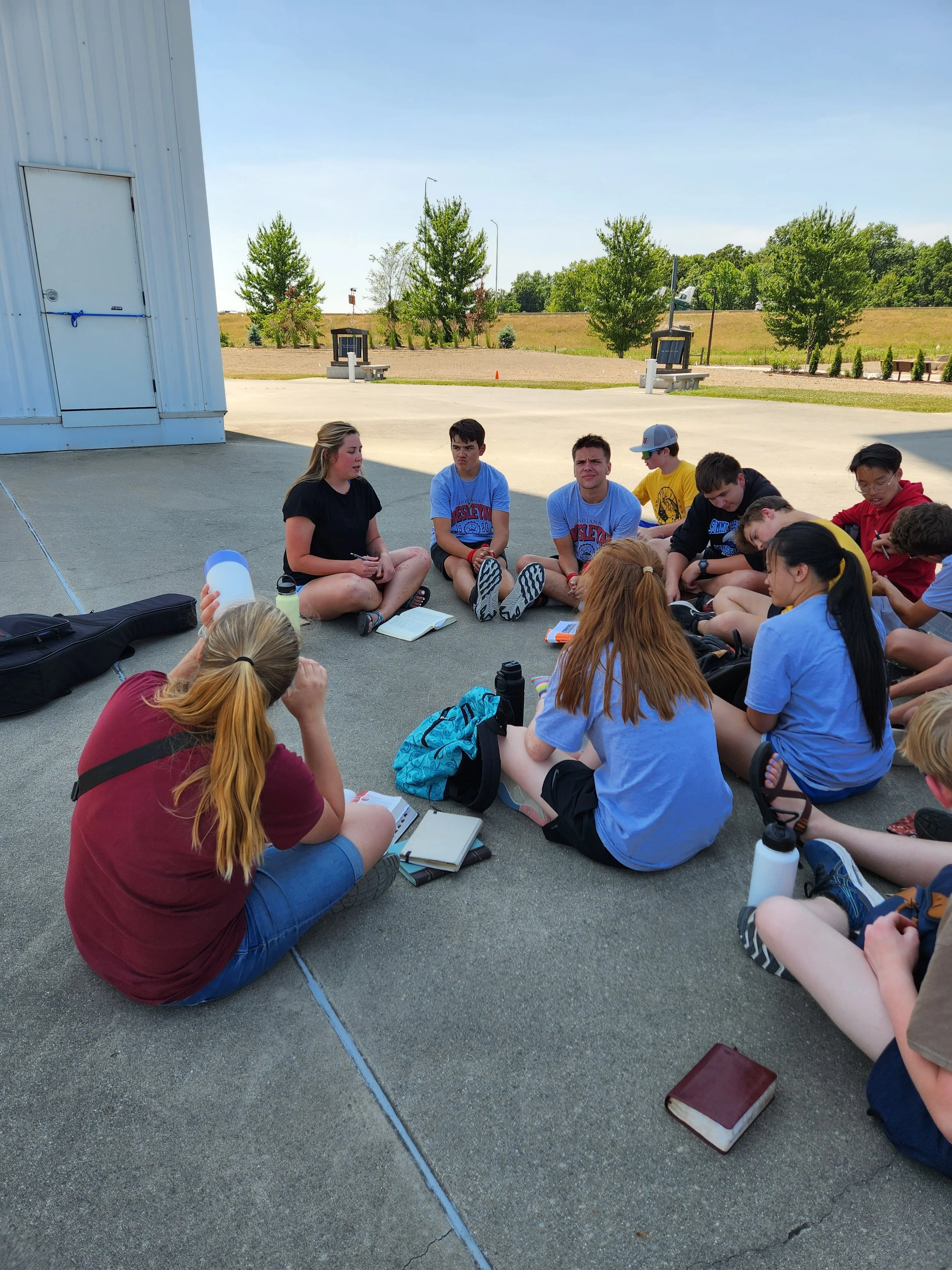 A group of teenagers sitting in a circle on the pavement outdoors, possibly participating in a discussion or group activity on a sunny day.