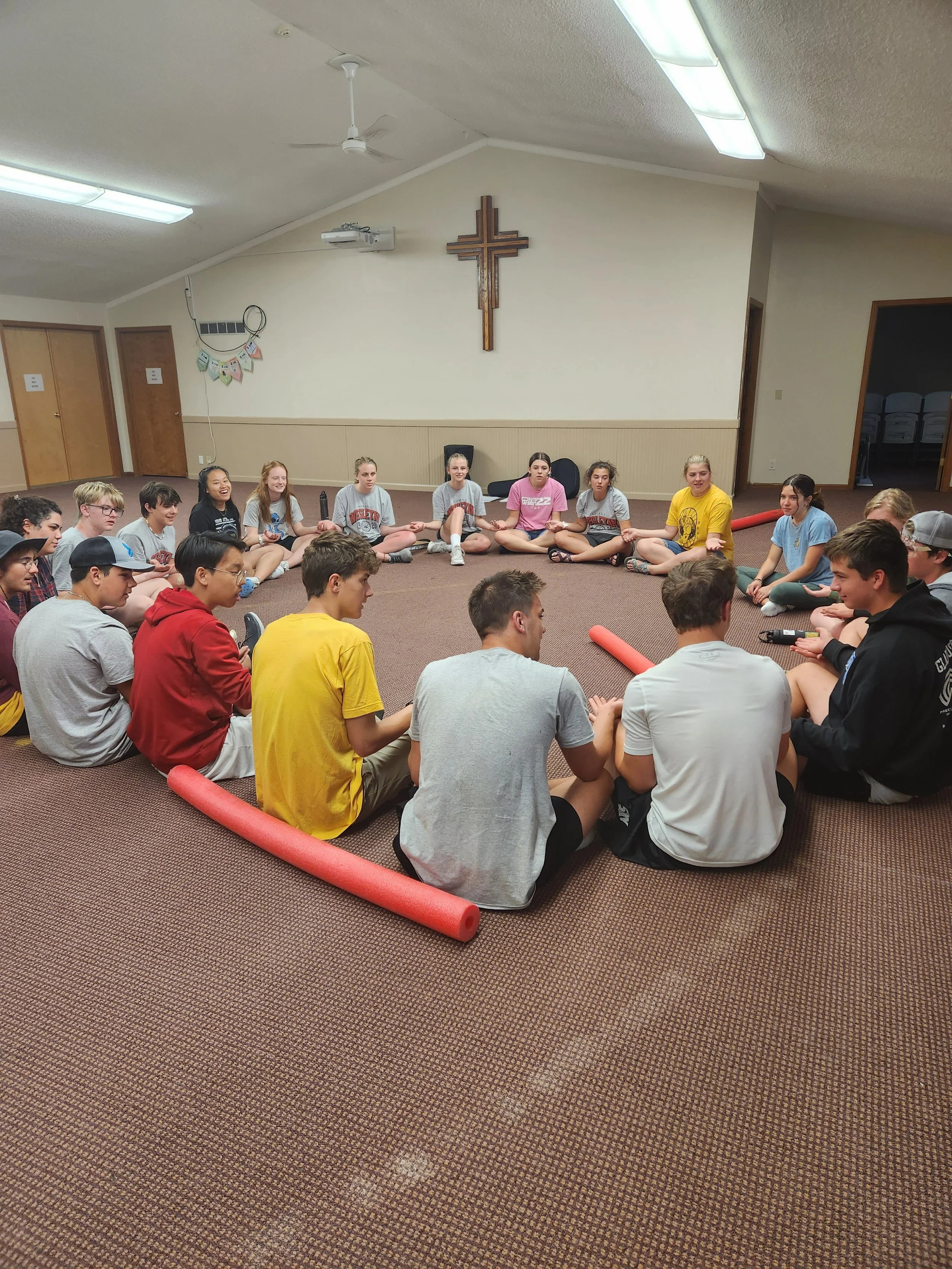 Group of teenagers sitting in a circle on a carpeted floor inside a church room with a wooden cross on the wall.