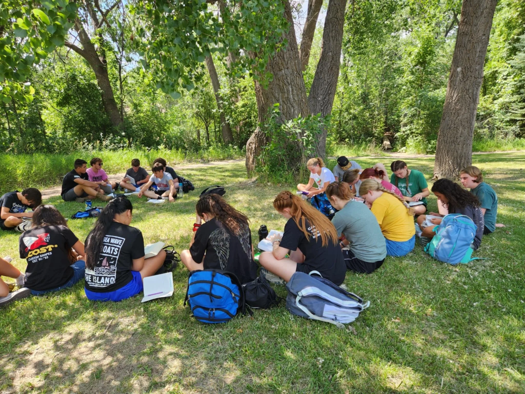 Group of teenage students sitting in a circle on grass in a shaded outdoor park, engaged in reading or writing activities.